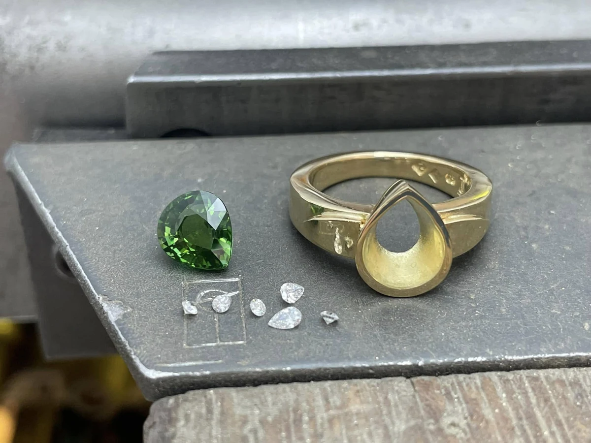 A gold ring setting with a green gemstone and loose clear stones on a jeweller’s workbench