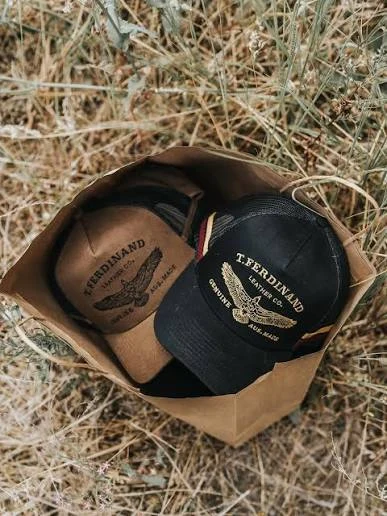 Brown and black T. Ferdinand Leather Co. caps in a paper bag on dry grass
